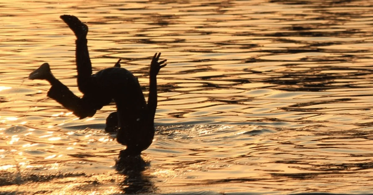 Silhueta de criança pulando na água ao pôr do sol, ilustrando cuidados com afogamento no verão